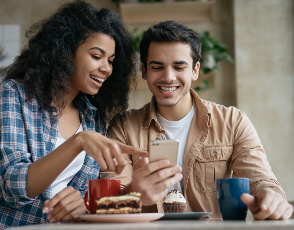 Un jeune couple consulte une annonce de location saisonnière sur leur smartphone, avec le sourire, autour d’un café et de pâtisseries. Une image qui illustre l’enthousiasme des vacanciers… mais qui rappelle aussi la nécessité de rester vigilant face aux arnaques de locations en ligne.